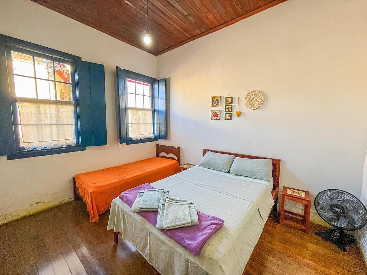 Quarto com cama de casal, cama de solteiro com colcha laranja e janelas azuis